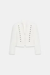 Blazer entallado color blanco de cuello redondo y manga larga con hombreras. Presenta detalles de aplicaciones de botones metálicos en el frente y forro interior combinado a tono.