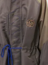 Campera tipo piloto con capucha, cierre frontal con botones y detalle de costuras en zigzag. Presenta cordones ajustables en la cintura y capucha, puños elásticos y logo bordado en el pecho.