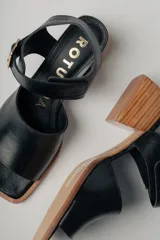 Sandalias de cuero negro con diseño minimalista y tacón ancho geométrico en tono madera. La capellada lisa aporta una estética depurada y moderna, mientras que la pulsera con hebilla metálica ajustable asegura un calce firme y confortable.