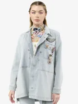Chaqueta de denim en tono azul claro con efecto lavado, corte holgado y cierre frontal con botones. Presenta múltiples insignias decorativas, incluyendo broches con cristales, una insignia con cinta y detalles de jacquard en los laterales. Cuenta con bolsillos frontales y aberturas laterales.