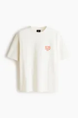 Remera de algodón blanco crudo, de corte oversized con hombros caídos y cuello redondo acanalado. Presenta pequeño estampado en el pecho con la inscripción "nas ©1994" en tono anaranjado.