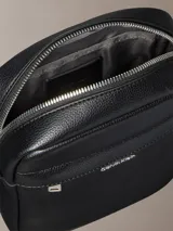 Cartera reporter estructurada Calvin Klein de poliuretano con textura granulada, color negro, cierre superior de cremallera y bolsillo frontal con cierre. Cuenta con correa de hombro ajustable y logo metálico de la marca en la parte delantera.