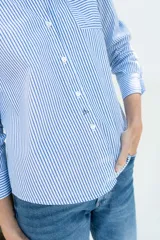 Camisa de popelina a rayas celestes y blancas, de estilo clásico, con cuello camisero, manga larga con puños ajustados, bolsillo frontal aplicado y botones de nácar.