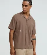 Camisa de manga corta, color marrón topo, con cuello camisero y corte amplio (boxy). Presenta bordados minimalistas en blanco en el frente, simulando un paisaje.