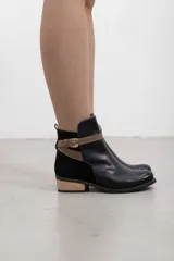 Botín de inspiración ecuestre de cuero liso negro combinado con gamuza, con correa marrón de cuero en contraste con hebilla metálica, cierre lateral interno y tacón bajo de madera.