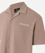 Camisa de manga corta, color marrón topo, con cuello camisero y corte amplio (boxy). Presenta bordados minimalistas en blanco en el frente, simulando un paisaje.