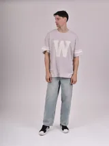 Remera gris de corte oversize con cuello redondo, mangas cortas con doble raya blanca y estampa frontal de una letra "W" grande en color blanco con efecto desgastado.
