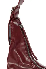 Cartera tipo hobo de material sintético con acabado brillante, cierre superior con cremallera, correa ajustable y laterales con cordones decorativos.