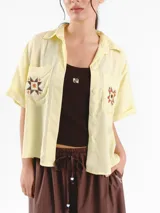 Camisa de manga corta color amarillo claro, con calce oversized, hombros caídos y cierre frontal con botones. Cuenta con bolsillos delanteros con bordados geométricos y mangas con dobladillo.