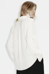 Camisa de manga larga color blanco hueso, con cuello camisero clásico y cierre frontal de botones. Presenta un corte recto y un bajo redondeado.