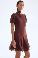 Vestido corto color marrón oscuro, con cuello alto y lazo frontal. Presenta mangas cortas abullonadas y una falda con vuelo y pliegues.