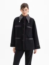 Chaqueta negra de corte recto con largo hasta la segunda cadera. Presenta detalles de cuero en cuello, frente, hombros y puños, con cuello decorado con tachas metálicas y punteras. Cuenta con bolsillos frontales tipo cargo.