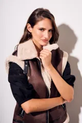 Chaleco oversize estilo gamulán en color marrón, confeccionado en cuero vegano con forro y solapas de corderito sintético en contraste. Posee cierre frontal metálico, bolsillos con cremallera y detalles de correas con hebillas en el cuello.