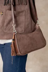 Cartera tipo baguette de diseño minimalista, confeccionada en una combinación de gamuza color beige en el panel frontal y cuero color chocolate en los bordes y la correa. Incluye un detalle colgante de cuentas de madera y cierre superior.