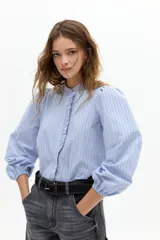 Camisa de algodón con estampado de rayas verticales en tonos azul y blanco. Presenta cuello mao, cierre frontal oculto con detalle de volado lateral y mangas abullonadas con puños elásticos.
