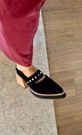 Zapatos tipo zueco de gamuza negra con punta fina y tira sobre el empeine decorada con tachas metálicas plateadas. Posee taco cuadrado de madera y suela con plataforma.
