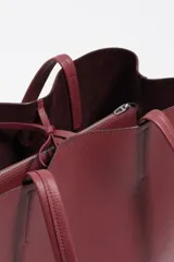 Bolso shopper de piel color borgoña, con textura granulada, asas de hombro y cierre de cordón. Presenta el logo metálico de Bimba y Lola en el frente.