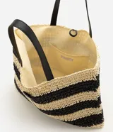Cartera tipo saco shopper confeccionada en paja trenzada, con diseño de rayas en color natural y negro. Cuenta con asas de hombro de material sintético y cierre con cordón ajustable con detalle de concha.