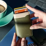Billetera tarjetero automática con estructura de aluminio y revestimiento de cuero bicolor en tonos verde oliva y beige. Cuenta con un cierre de broche a presión y el logo de la marca grabado en la parte inferior.