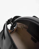 Cartera de mano color negro de cuero sintético, modelo Fiala de XL Extra Large, con diseño estructurado, asa superior y detalles de costuras y tiras decorativas laterales con hebilla. Incluye el logo metálico de la marca en el frente.