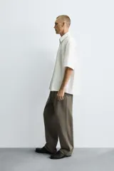 Camisa de algodón blanca con finas rayas verticales negras, corte relajado (relaxed fit) y manga corta. Presenta un bordado en hilo rojo en el lado izquierdo del pecho.
