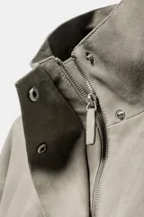 Chaqueta corta de ante con cuello alto, manga larga y cierre frontal con cremallera y botones a presión. Presenta bolsillos frontales con solapa y bajo abullonado con pliegues.