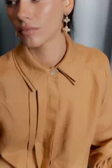 Camisa de lino color tostado con diseño de doble cuello, cartera escondida y tablas frontales que aportan movimiento. Presenta mangas largas y un corte versátil.