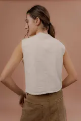 Chaleco color crudo sin mangas, con escote en V y bordados artesanales florales en tono amarillo, rosa y marrón en el frente. Presenta detalles de pasamanería con pompones pequeños en los bordes.