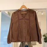 Camisa marrón con cuello en V, detalles bordados en los hombros y aplicaciones de encaje y alforzas en el frente. Tiene cierre frontal de botones y mangas largas abullonadas con puños ajustados.