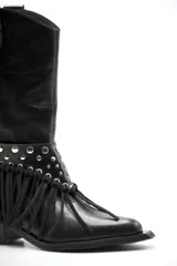 Bota de cuero negro estilo texano, con taco bajo y punta fina. Incluye un aplique desmontable en la zona del empeine decorado con tachas metálicas y flecos largos.