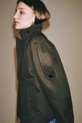 Trench corto Zara Woman Collection confeccionado en hilatura con algodón, en color verde militar. Presenta cuello solapa, manga larga con trabillas y botones en los puños, bolsillos delanteros, bajo asimétrico y cierre frontal cruzado con botones.