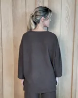 Remerón de textura acanalada en color gris claro, con cuello redondo, mangas largas y bolsillo frontal en el pecho. Presenta un diseño holgado con corte asimétrico, siendo más largo en la parte posterior.