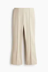 Pantalón tobillero color beige, de tiro alto con elástico oculto en la cintura y cierre lateral oculto. Piernas ajustadas con pliegues cosidos adelante y bajo acampanado.