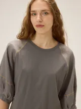 Remera gris oscuro de cuello redondo, confeccionada en algodón con spandex, con mangas raglán amplias y costuras pespunteadas en hilo contrastante de color ocre.