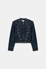 Chaqueta de denim de color azul oscuro, con cuello redondo, hombreras y cierre frontal de cremallera. Presenta diseño de inspiración militar con apliques de botones metálicos en la parte delantera y bajo elástico.