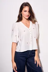 Blusa blanca con delicados bordados pequeños en contraste, escote en V y cierre frontal con botones. Mangas cortas abullonadas y corte fluido.