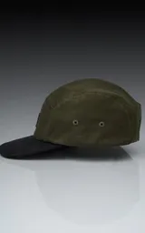 Gorra estilo 5 paneles, color verde militar en la corona y visera plana negra. Presenta un pequeño parche cuadrado negro con un logo blanco bordado en forma de tijeras o lazo.