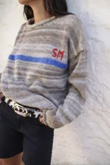 Sweater de punto en tonos beige con efecto degradado, cuello redondo y manga larga. Presenta una franja horizontal en color azul y el bordado de las letras 'SM' en color rojo en el pecho.