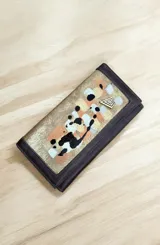 Billetera grande de cuero marrón, con solapa frontal decorada con detalles pintados a mano en tonos dorados, blancos, negros y terracota, y aplique metálico triangular de la marca.