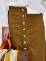 Pollera midi de denim color marrón, de tiro medio y corte recto. Presenta cierre frontal con botones metálicos a la vista y un tajo central en el ruedo con terminación deshilachada.