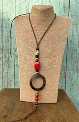 Collar largo con tira de cuero marrón, decorado con abalorios de madera, resina roja y blanca, y un gran aro de madera en el centro.