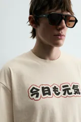 Camiseta relaxed fit color crema, con cuello redondo y manga corta. Presenta un estampado frontal con caracteres japoneses en negro y contorno rojo.