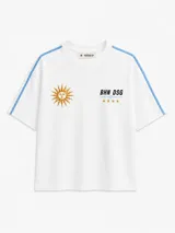 Remera blanca de algodón, de corte oversize y manga corta, con bandas celestes en los hombros y mangas. Presenta un sol bordado en el pecho derecho y la inscripción "BHN DSG" junto a cuatro estrellas bordadas en el pecho izquierdo.