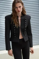 Blazer corta de estilo militar en color negro, con cuello redondo, manga larga y hombreras marcadas. Presenta un diseño frontal con bordados decorativos a tono y botones dorados, cierre con gancho metálico y forro interior.