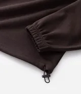 Buzo de corte holgado tipo ballon, color marrón oscuro, con cuello alto y cierre frontal parcial. Presenta mangas largas con puños elásticos y cordón ajustable en el ruedo.