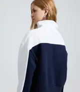 Buzo tipo polo de textura waffle con diseño color block en blanco y azul marino. Presenta cuello alto con cierre de botones, mangas largas y bordados decorativos en el frente.
