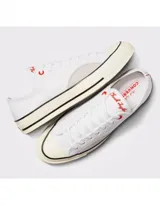 Championes Converse modelo Chuck 70 Ox, de corte bajo, confeccionados en lona blanca con detalles en gamuza gris claro en los laterales. Presentan suela de goma con franjas negras y ojal superior en color rojo.