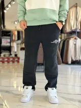 Pantalón de jogging oversize negro con felpa, de corte recto y cordón ajustable en la cintura. Presenta un pequeño estampado de texto blanco en el lateral izquierdo.