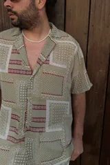 Camisa de manga corta con cuello clásico y estampado geométrico en tonos verde, beige y rojo. Cierre frontal con botones. Corte recto.