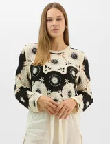 Buzo de tejido crochet con diseño de motivos circulares en contraste blanco y negro, con apliques de pedrería en el centro de los círculos. Presenta corte recto, cuello redondo y manga larga.
